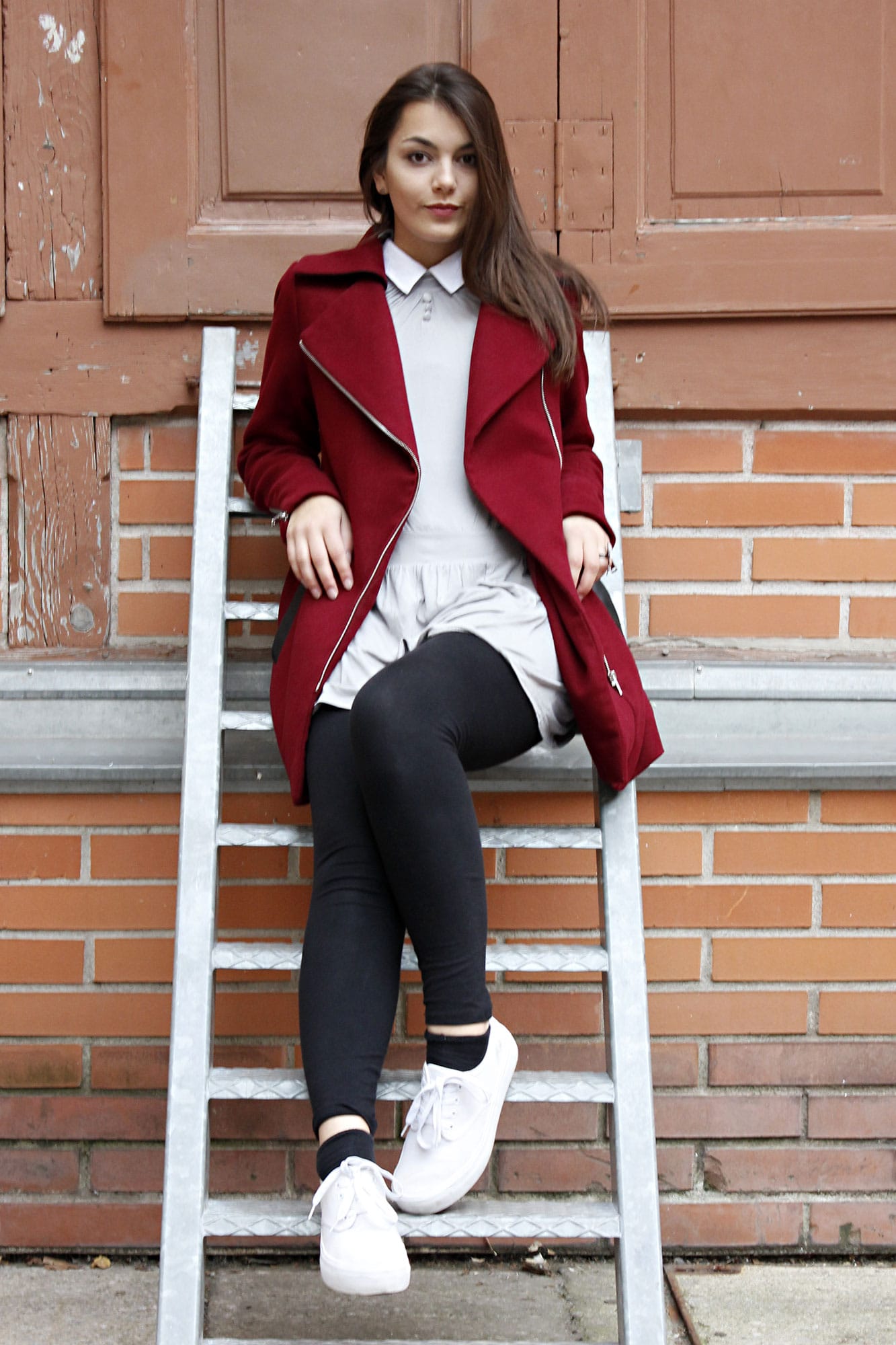 Dorie is sitting on a ladder, wearing a red coat, black legging, a grey dress and white shoes 