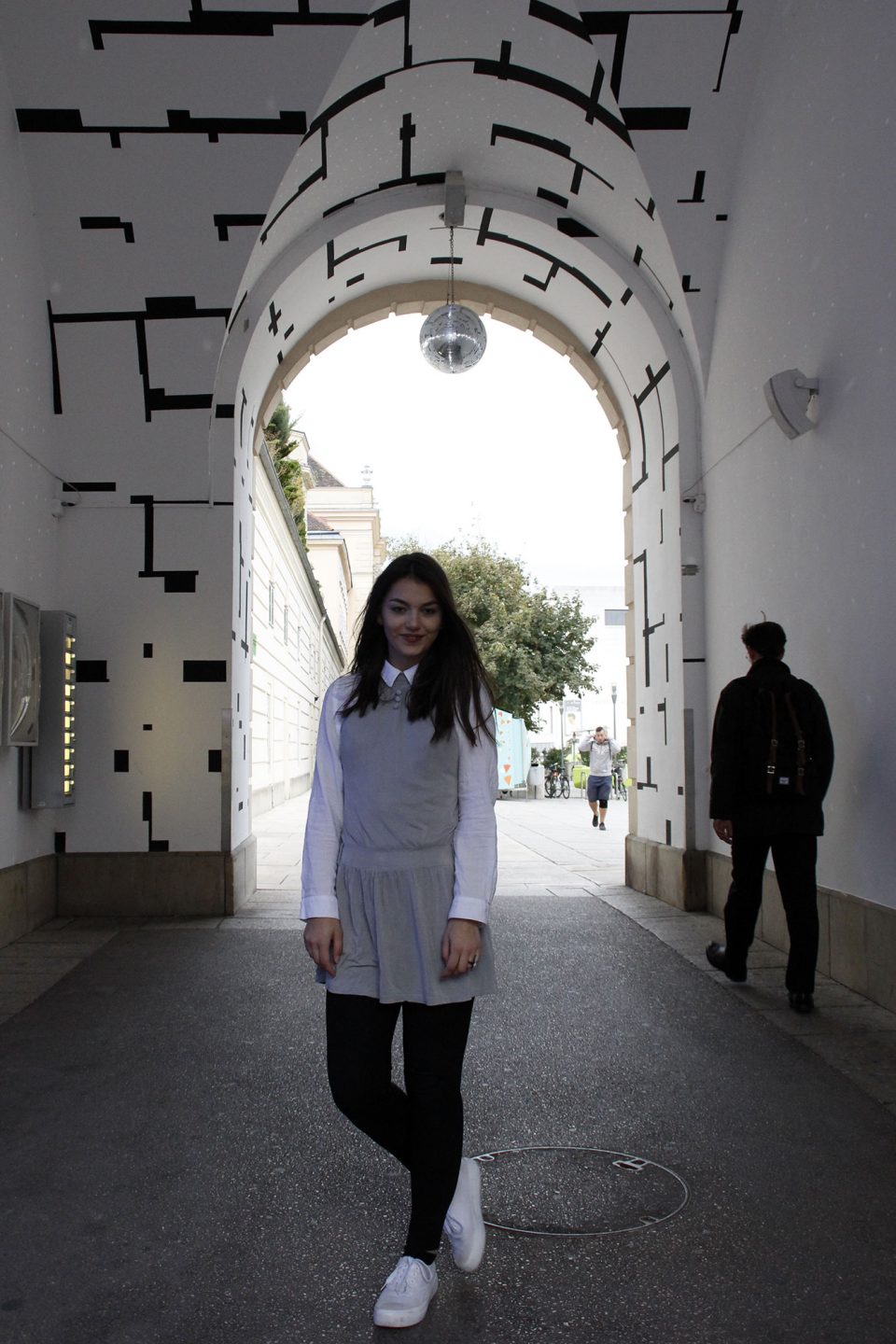 Dorie standing in a black white tunnel, waring a grey dress, a white blouse, black legging and white shoes
