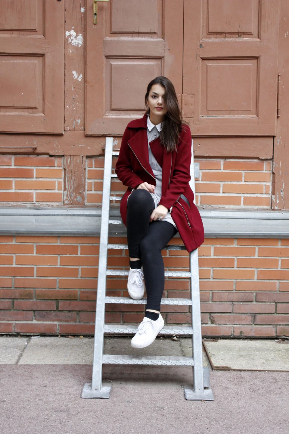Dorie sitting on a ladder, wearing a red coat from Stradivarius, a grey dress, black legging and white shoes from even&odd