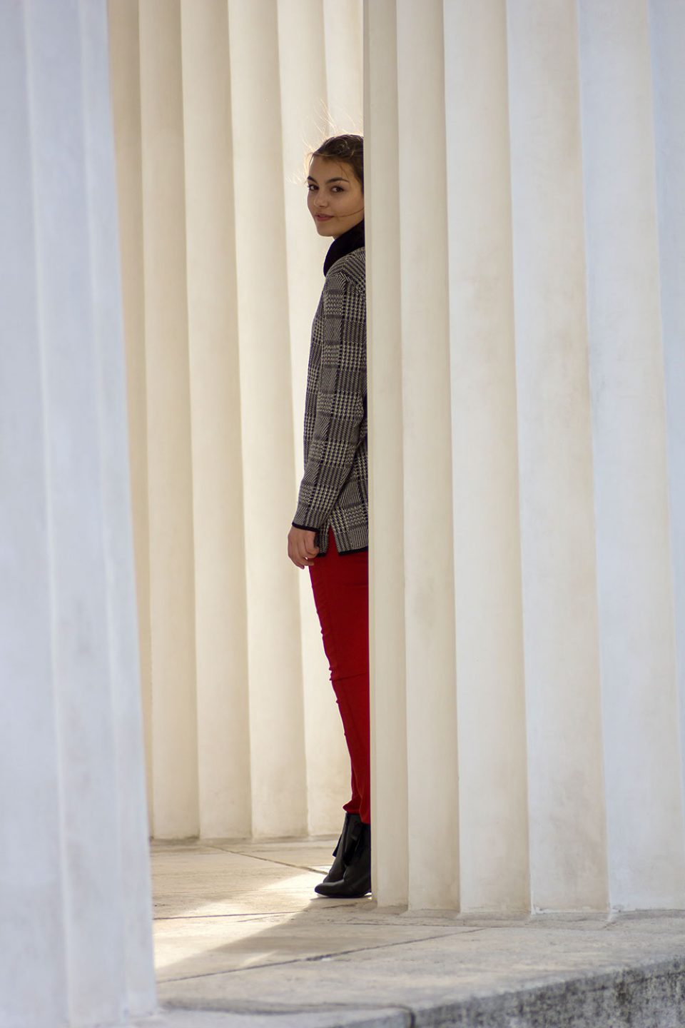 Dorie steht hinter einer Säule, trägt eine rote Hose vonl H&M, schwarze Heels von Mint&Berry und einen grauen Pullover