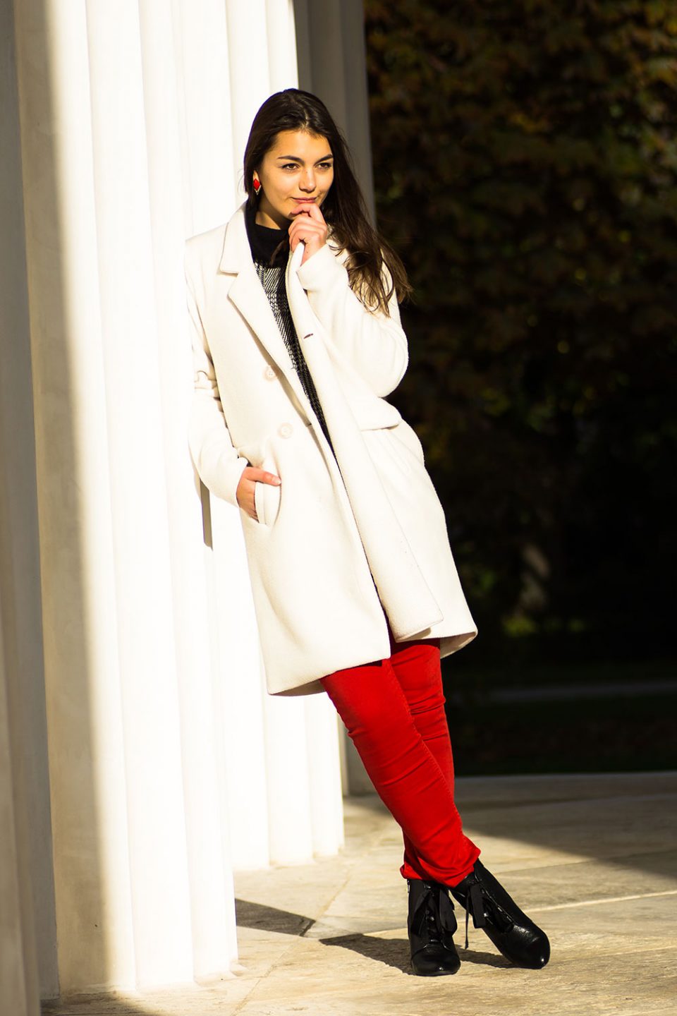 Dorie lehnt an einer Säule, trägt einen weißen Mantel, eine rote Hose von H&M, schwarze Stiefeletten von Mint&Berry und einen grauen Pullover